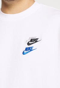 Vit bomulls t-shirt med tre broderade Nike-loggor i svart, grått och blått. Klassisk crew neck-design med korta ärmar.