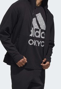 Svart idrottsjacka med dragkedja fram, texturerad tyg och en svart t-shirt med en vit grafisk logotyp. Bärs med svarta byxor.