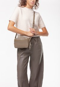 Piccolo borsa a tracolla verde oliva con un pannello in tessuto texturizzato, hardware argentato e una tracolla regolabile, tenuta da una persona che indossa un maglione chiaro.
