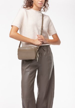 Piccolo borsa a tracolla verde oliva con un pannello in tessuto texturizzato, hardware argentato e una tracolla regolabile, tenuta da una persona che indossa un maglione chiaro.