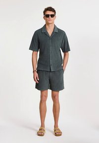 Sötétzöld rövid ujjú, gombos ing texturált anyagból, hozzá passzoló rövidnadrággal és barna szandálokkal. Napszemüveg viselve.