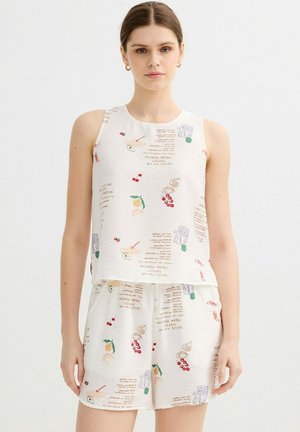 Joven mujer con top blanco sin mangas y pantalones cortos con estampados coloridos de frutas y texto, de pie contra un fondo liso.