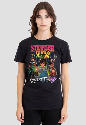 Young woman wearing black Stranger Things "Tales from the 85" t-shirt featuring illustrated characters and text "We Are The" on front.