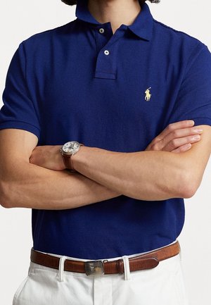 Mężczyzna w granatowej koszulce polo z żółtym logo konia, białe spodnie, brązowy skórzany pasek i zegarek na nadgarstek z brązowym paskiem, skrzyżowane ręce.