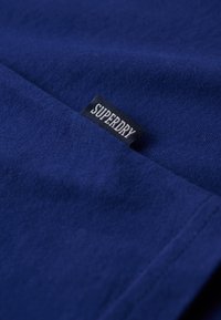 Camiseta de algodón azul marino con una textura suave, que presenta una pequeña etiqueta negra con la inscripción "SUPERDRY" cerca de la costura.