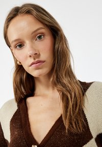 Braun- und cremefarbener Cardigan mit V-Ausschnitt, der mit einem strukturierten Strick und einem Rautenmuster ausgestattet ist. Akzentuiert mit einem Knopf in Goldoptik.