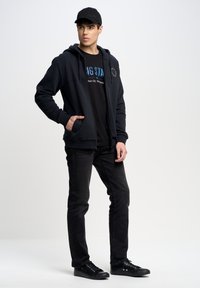 Sudadera negra con cremallera y bolsillos frontales, superpuesta a una camiseta gráfica negra. Combinada con jeans negros y zapatillas negras con cordones. Gorra negra.