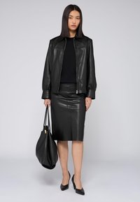 Černá kožená bunda se zipem vpředu, spárovaná s černou koženou tužkovou sukní. Doplňky zahrnují velkou černou tašku a špičaté podpatky.