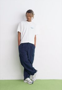 Tricou alb cu logo albastru, pantaloni cargo bleumarin și adidași albi. Stă sprijinit de un perete alb pe un pardoseală verde. Prezintă un croi relaxat.