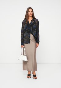 Blusa nera trasparente con motivi floreali blu e grigi, abbinata a una gonna maxi beige, sandali neri e una borsa bianca.