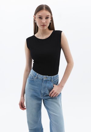 Jeune femme aux cheveux longs portant un haut sans manches noir, un jean taille haute bleu clair et des boucles d'oreilles argentées en forme d'anneau, posant devant un fond blanc.