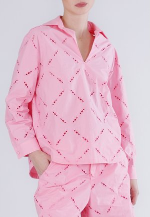 Mujer vistiendo un conjunto rosa a juego con patrones de recortes en forma de diamante, mangas largas y una camiseta con escote en V, manos en los bolsillos del pantalón.