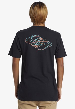 Persona con capelli biondi corti che indossa una t-shirt nera con un disegno astratto blu e arancione sulla schiena e pantaloni beige.