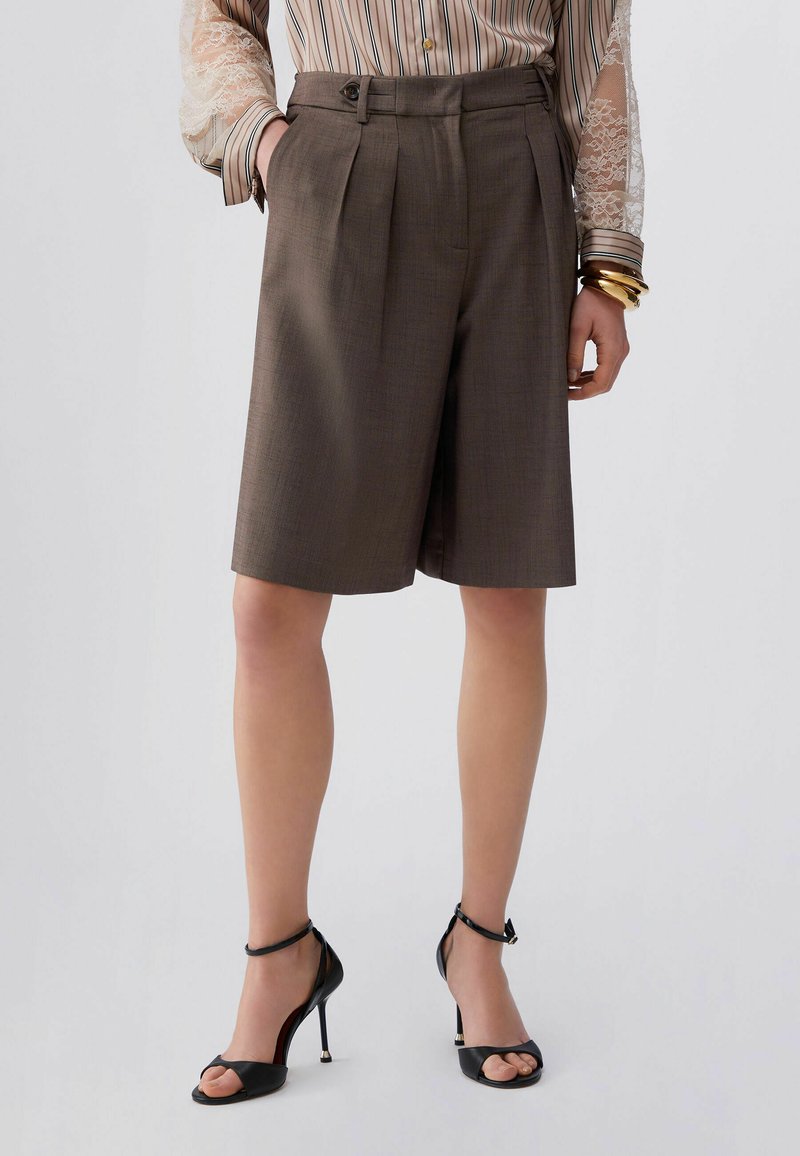 Shorts marron sur mesure avec des plis, dotés de poches latérales et d'une texture lisse. Associés à des talons noirs à brides et un chemisier rayé.