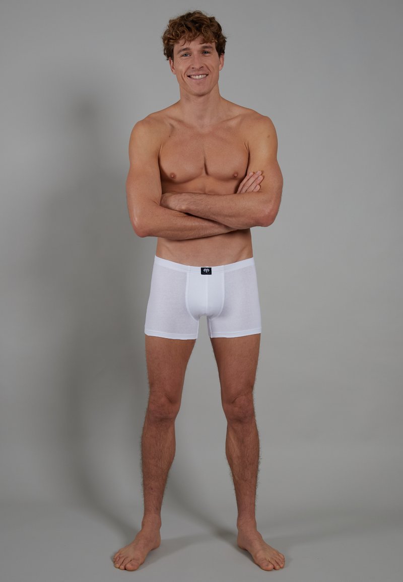 Weiße Baumwoll-Boxershorts mit weicher Textur und einem kontrastierenden Taillenbund-Logo, an einem Modell präsentiert. Neutraler grauer Hintergrund.