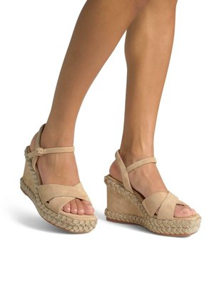Beige Keilsandalen mit überkreuzten Wildlederriemen und Knöchelriemen. Merkmale eine gewobene Jute-Zwischensohle mit einem dekorativen metallischen Akzent.