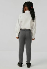 Vit skjorta med volangdetaljer på axlarna, grå skräddarsydda byxor och svarta sneakers. Skjortan har långa ärmar och knäppta manschetter.