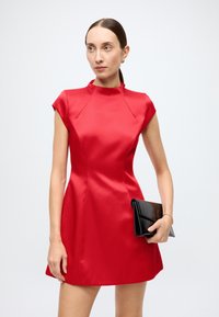 Robe en satin rouge avec un col montant et des manches courtes, présentant une silhouette ajustée. Modèle tenant une pochette noire.