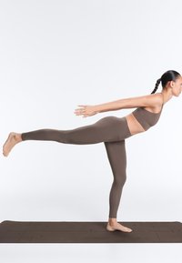 Vrouw in bruine sportkleding die op één been balanceert in de yoga Warrior III-houding op een donker yogamat tegen een witte achtergrond.