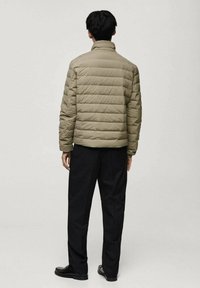 Veste matelassée en vert olive, design matelassé avec des lignes horizontales, col haut, associée à un pantalon et des chaussures noirs. Vue de dos.