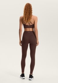 Smeđe sportski grudnjak s ukriženim naramenicama i odgovarajućim visokim tajicama od rastezljivog materijala. Na pojasu se nalazi logo.