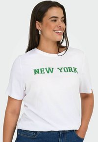 Šypsosi moteris su ilgais tamsiais plaukais, vilkinti sidabriniais auskarais, balta marškinėliais su žaliais užrašais "NEW YORK" ir mėlynomis džinsomis, viena ranka kišenėje.