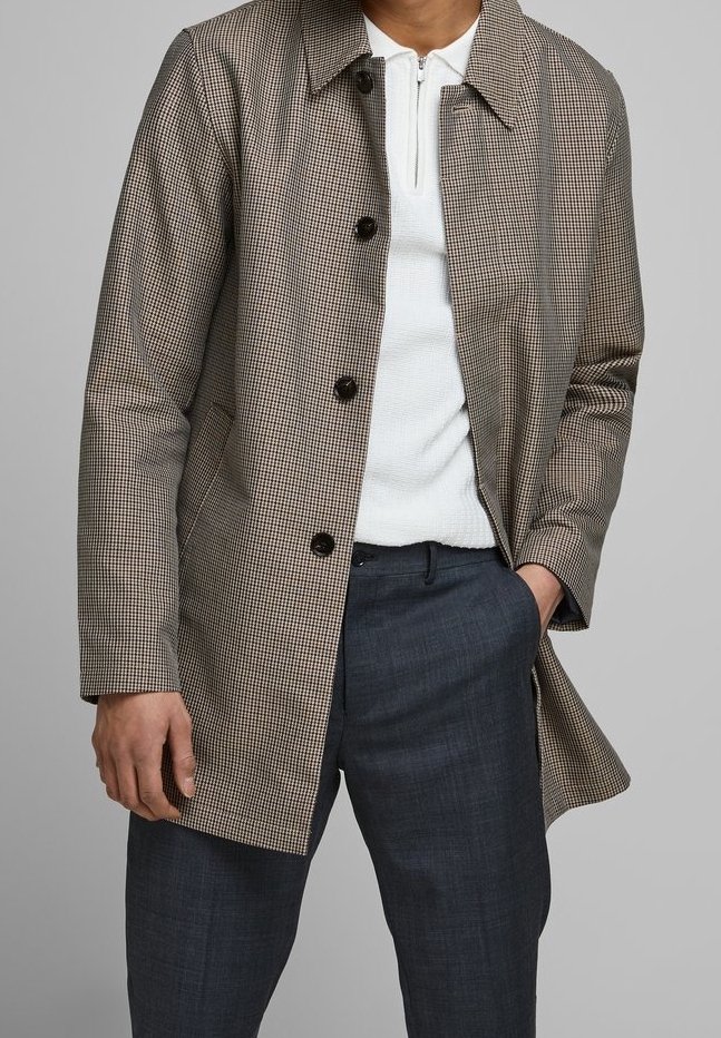 Muž má na sobě hnědý kostkovaný kabát přes bílou svetr s zipem a tmavě šedé kalhoty, s jednou rukou v kapse na šedém pozadí.
