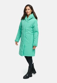 Mintgroene gewatteerde jas met een omvouwbare capuchon, vooraan met drukknoopsluiting, twee zijzakken en een middellange snit, gedragen over zwarte broeken en enkellaarsjes.