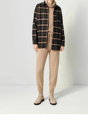 Person, der bærer beige rullekrave sweater og bukser med bindebånd i taljen, brune snørebåndsstøvler og sort-brun ternet jakke, står imod en ensfarvet væg.