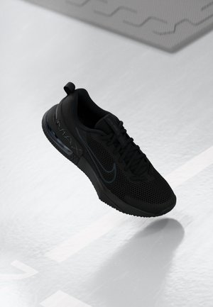 Baskets Nike Air Max noires avec tige en mesh, semelle à coussin d'air visible et lacets, présentées suspendues au-dessus d'un sol clair avec des motifs géométriques.