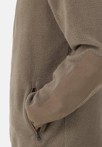 Close-up van een persoon die een bruine fleecjas met een suède elleboogpatch draagt, met de hand in de ritszak aan de zijkant gestoken.