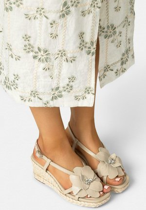 Sandalias de cuña beige con grandes decoraciones florales y centros de piedras de imitación, usadas con un vestido crema bordado con flores verdes.