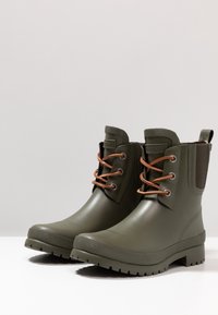 Olivově zelené gumové boty do kotníků s hnědými tkaničkami, bočními elastickými panely, texturovanou podrážkou a plochým podpatkem pro stabilitu.