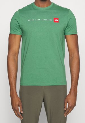 Mężczyzna w zielonym T-shircie z krótkim rękawem z napisem "Never Stop Exploring" i czerwonym logo The North Face na piersi, w zielonych spodniach.
