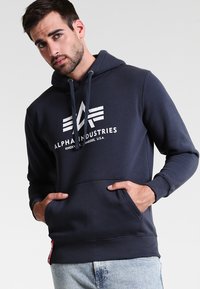 Marineblauwe hoodie van zachte stof. Beschikt over een voorzak, elastische manchetten en een groot wit logo op de borst.