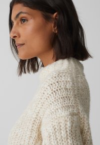 Maglione a maglia color crema con un colletto alto e un modello testurizzato, caratterizzato da una vestibilità ampia e dettagli sottili nella scollatura.