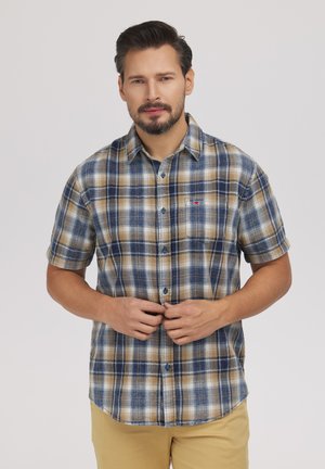 Mężczyzna zapinający guziki na niebiesko-beżowo-białej kratkowanej koszuli z krótkim rękawem, noszący beżowe spodnie, na jednolitym jasnoszarym tle.