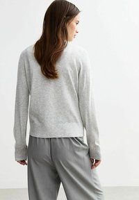 Donna con i capelli lunghi e castani, che indossa un maglione grigio chiaro e pantaloni grigi larghi, di spalle contro uno sfondo bianco semplice.