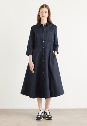 Navy jurk met knoopsluiting aan de voorkant, lange mouwen, een borstzak en een uitlopende rok. Gecombineerd met sportieve witte en navy sneakers.