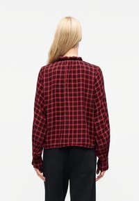 Ξανθό άτομο που έχει την πλάτη γυρισμένη, φοράει κόκκινο και μαύρο καρό πουκάμισο με μακριά μανίκια, γιακά με βολάν και μαύρο παντελόνι, σε λευκό φόντο.