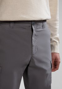 Nahaufnahme von grauen Hosen mit geknöpftem Bund und Reißverschluss, getragen mit einem beigen Langarmoberteil, Hand ruht nahe der Seitentasche.