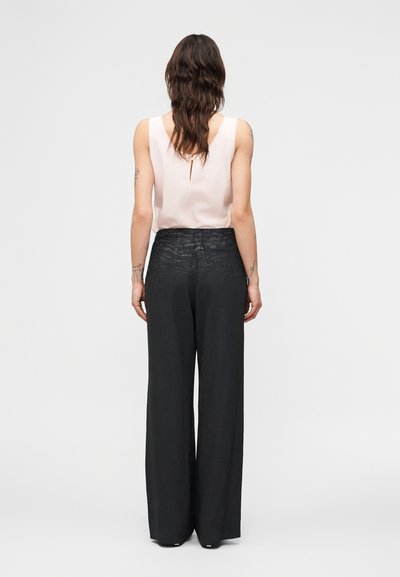 Γυναίκα με μακριά κυματιστά μαλλιά που φορά ανοιχτό ροζ μπλουζάκι χωρίς μανίκια και ψηλόμεση φαρδιά μαύρη παντελόνα, στέκεται γυρισμένη με την πλάτη σε απλό φόντο.