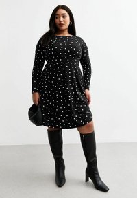 Robe noire à manches longues avec des pois blancs, corsage ajusté, jupe évasée, portée avec des bottes noires à talons montant jusqu'aux genoux et un sac à main noir.