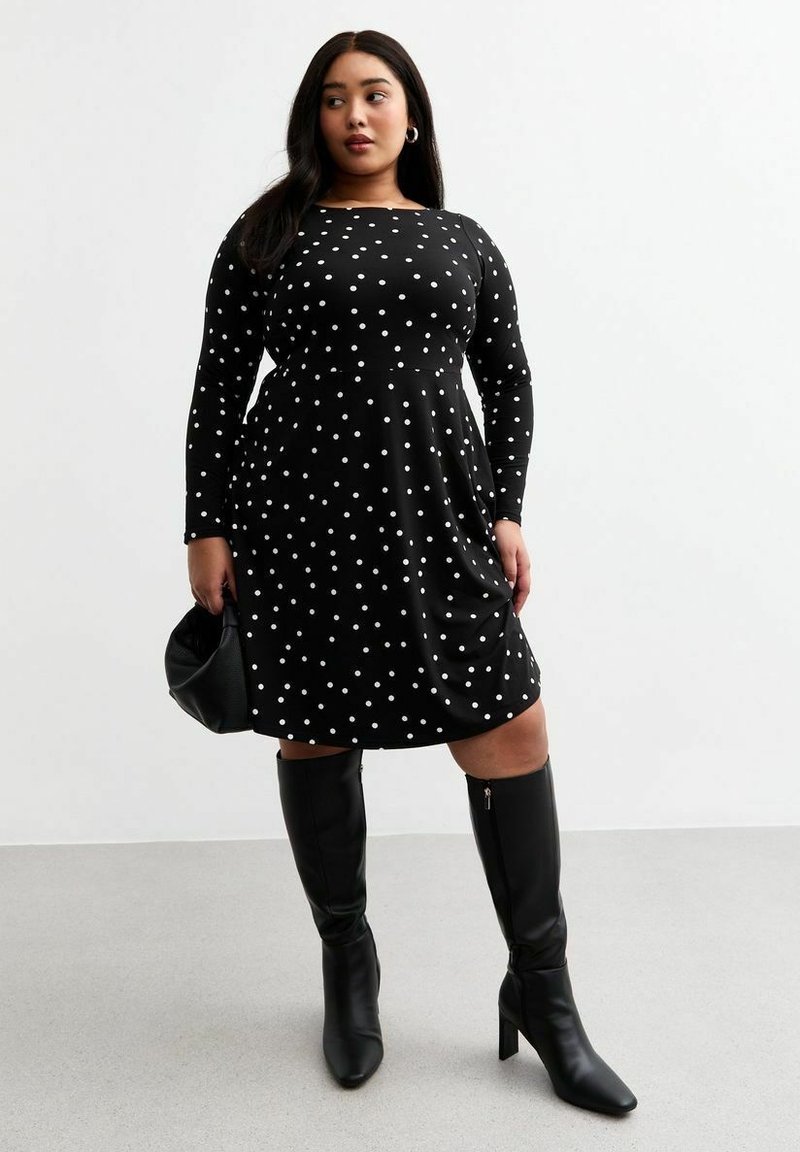 Robe noire à manches longues avec des pois blancs, corsage ajusté, jupe évasée, portée avec des bottes noires à talons montant jusqu'aux genoux et un sac à main noir.