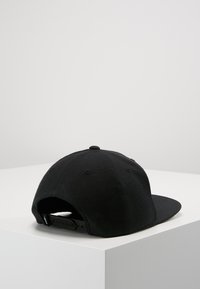Cappellino snapback nero regolabile poggiato su una superficie bianca con angoli su uno sfondo grigio chiaro uniforme.