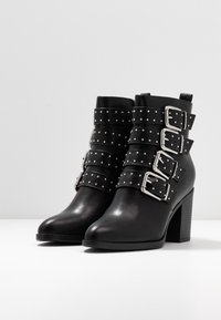 Botas negras hasta el tobillo con punta afilada, que presentan múltiples correas anchas decoradas con tachuelas y hebillas plateadas, y un tacón grueso.