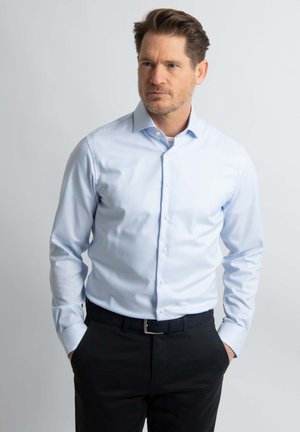 Lichtblauw overhemd met knopen in een soepele stof, lange mouwen en een klassieke kraag; gedragen met donkere broek en een zwarte riem.