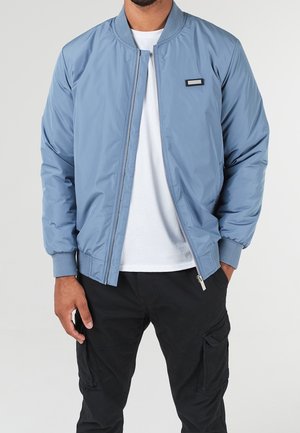 Man wearing light blue bomber jacket over white shirt and black cargo pants with one hand in pocket, against plain background.