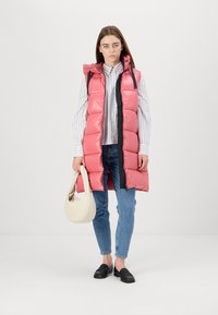 Gilet imbottito rosa con cappuccio; design trapuntato, zip frontale e dettagli neri. Abbinato a una maglietta a righe, jeans blu e una borsa a mano color crema chiaro.