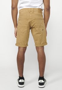Shorts en denim beige avec des ourlets retroussés, comportant deux poches arrière avec un design cousu. Portés avec des baskets noires et blanches.